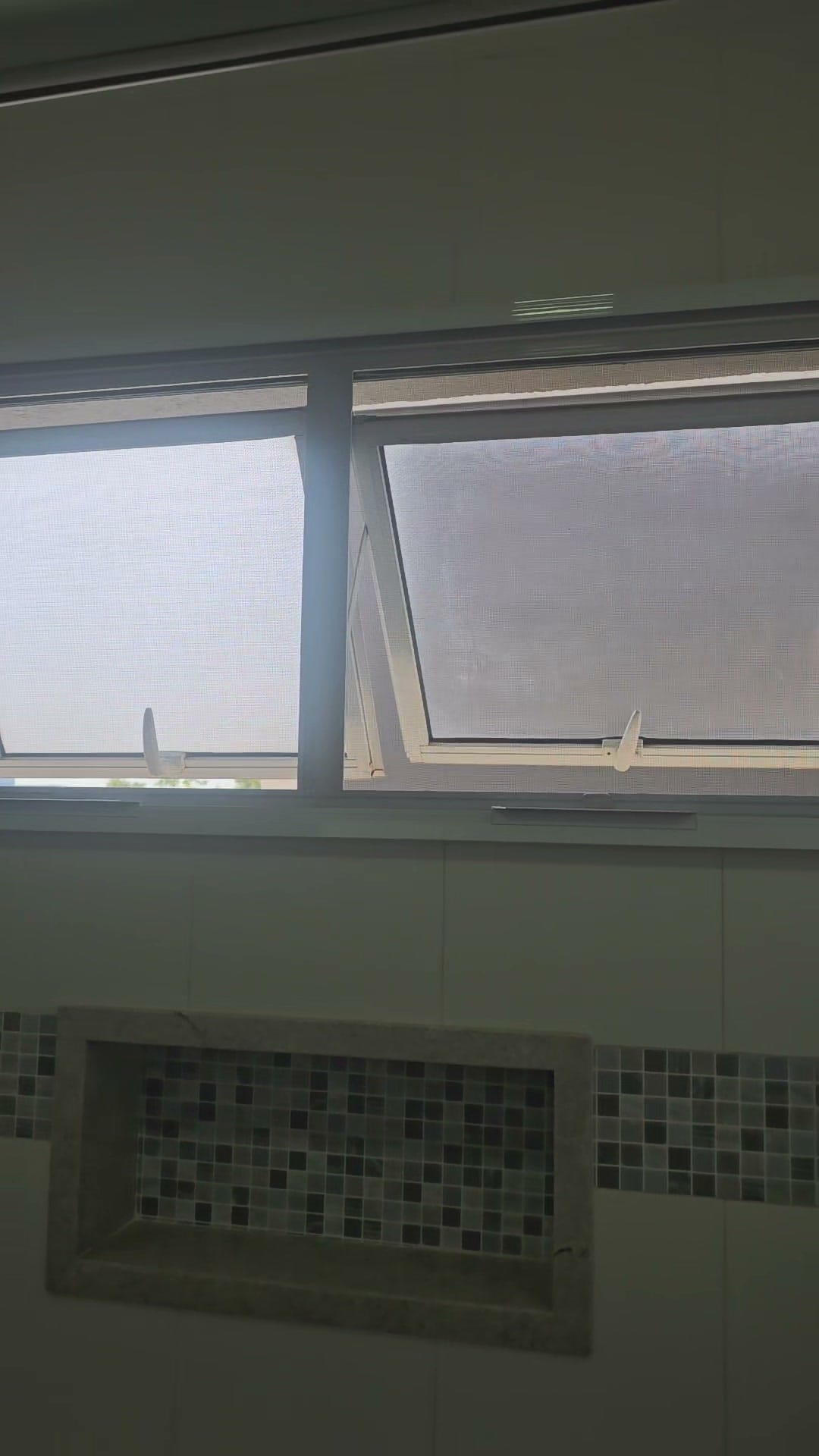 Duas janelas maxim-ar de alumínio branco com tela mosquiteira modelo guilhotina instalada, vistas de dentro do banheiro