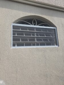 Janela em arco com grade decorativa de ferro e tela mosquiteira instalada por dentro do vão, em parede bege