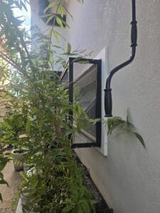 Janela maxim-ar com tela mosquiteira instalada, vista de fora, com plantas e ramos na frente da parede clara