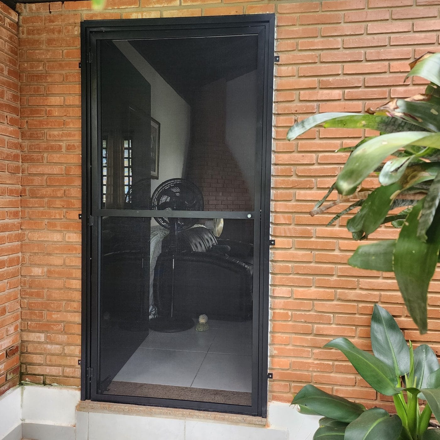 Porta de tela mosquiteira de quadro preto em parede de tijolo aparente em São Pedro, dando para a área da churrasqueira