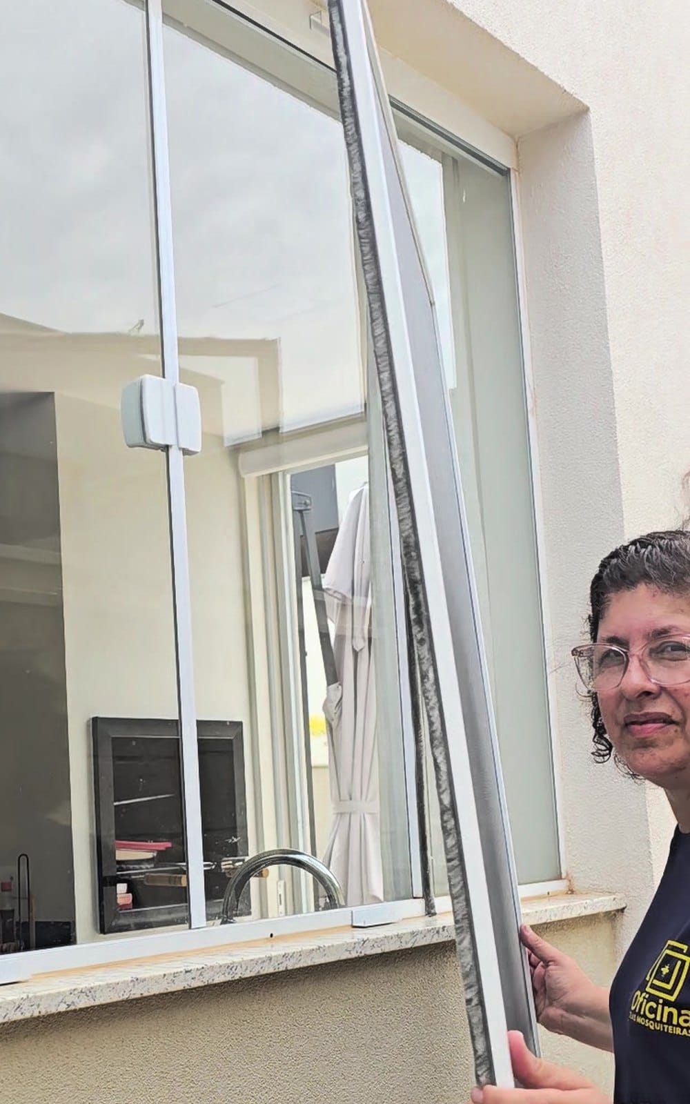 Andréa, da Oficina Telas Mosquiteiras, segurando uma tela mosquiteira removível retirada da janela; a escova de vedação cinza corre por todo o perfil de alumínio da peça
