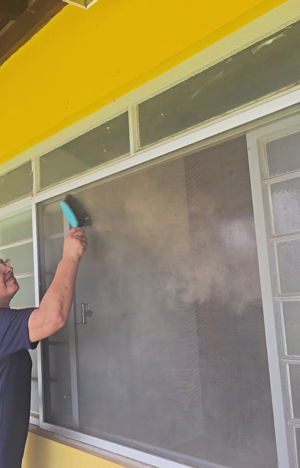 Andréa, da Oficina Telas Mosquiteiras, tirando a poeira da malha de uma tela mosquiteira de correr com movimento leve, de cima para baixo